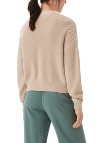 Beige gestrickter Pullover mit lockerer Passform, gerippten Bündchen und Saum, kombiniert mit grünen Hosen. Das strukturierte Material verleiht ein weiches Aussehen.