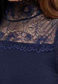 Top marine avec un col en dentelle haute, motifs floraux, texture transparente et bords en dentelle, contrasté avec une base en tissu uni.