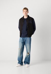 Black and navy two-tone jacket with snap buttons, paired with a dark blue sweatshirt and light blue ripped jeans, and white sneakers.