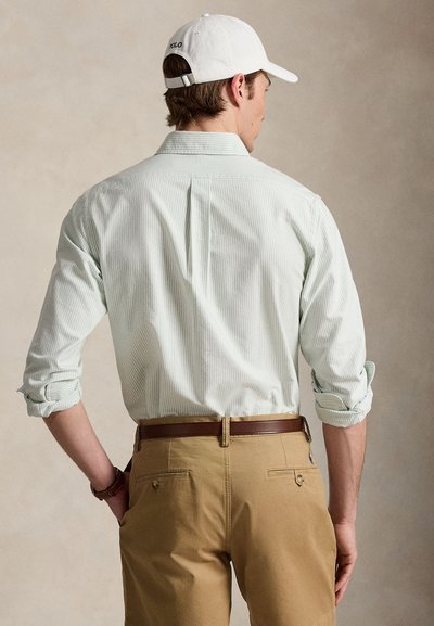 Homme portant une chemise claire à rayures avec les manches retroussées, pantalon beige, ceinture marron et casquette de baseball blanche, vu de dos sur un fond beige.