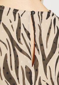 Kurzärmlige Bluse aus leichtem Chiffon. Beige mit schwarzen Zebra-Streifen und weißen Punktemustern. Verfügt über einen Knopfverschluss auf der Rückseite.