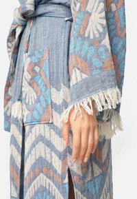 Blauwe en beige gewatteerde robe met geometrische ontwerpen, met franjes aan de mouwen en een voorzak, gemaakt van textiel met structuur. Hand op de zij rustend.