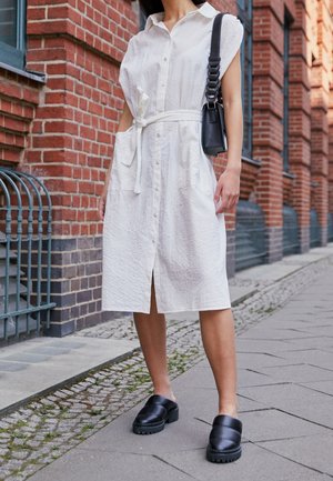 Vit, ärmlös skjortklänning med ett strukturerat mönster, cinchad midja och sidofickor. I kombination med svarta klumpiga slip-on skor.