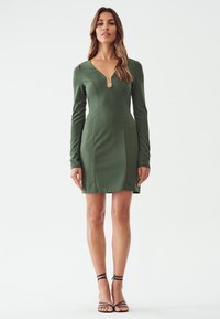 Robe verte à manches longues avec un décolleté en V et une découpe en forme de trou de serrure. Silhouette ajustée et texture lisse, accompagnée de sandales à talons noirs.