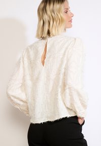 Blusa crema con tessuto texturizzato, maniche ampie e schiena aperta. Presenta un sottile motivo floreale e un orlo sfilacciato.