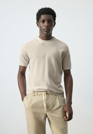 Ung mand med kort krøllet hår iført beige strikket kortærmet skjorte og lyse khakibukser, stående mod en ensfarvet lys baggrund.