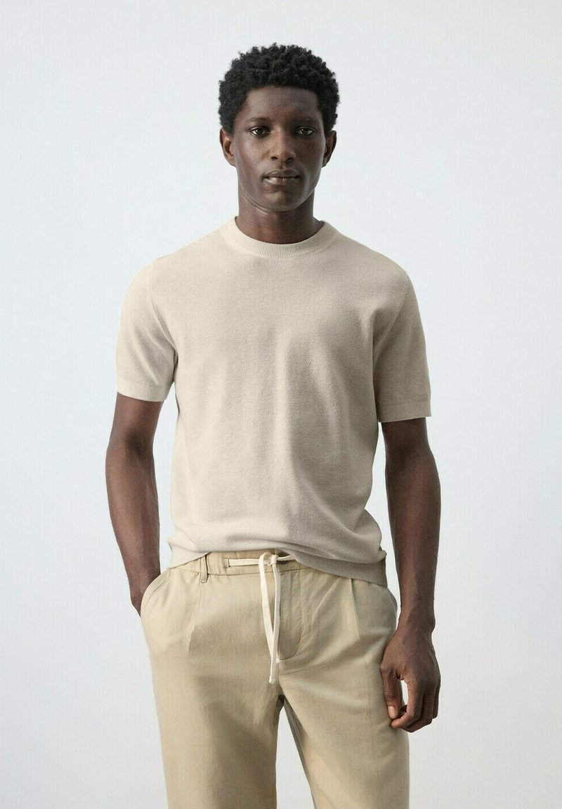 Jeune homme aux cheveux courts et bouclés portant un t-shirt en maille beige à manches courtes et un pantalon kaki clair, debout devant un fond clair uni.