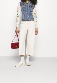 Džínová bunda s květinovou výšivkou, bílé pletené rukávy, bílé široké kalhoty, bílé tenisky a červená kabelka. Hladké textury po celém outfitu.