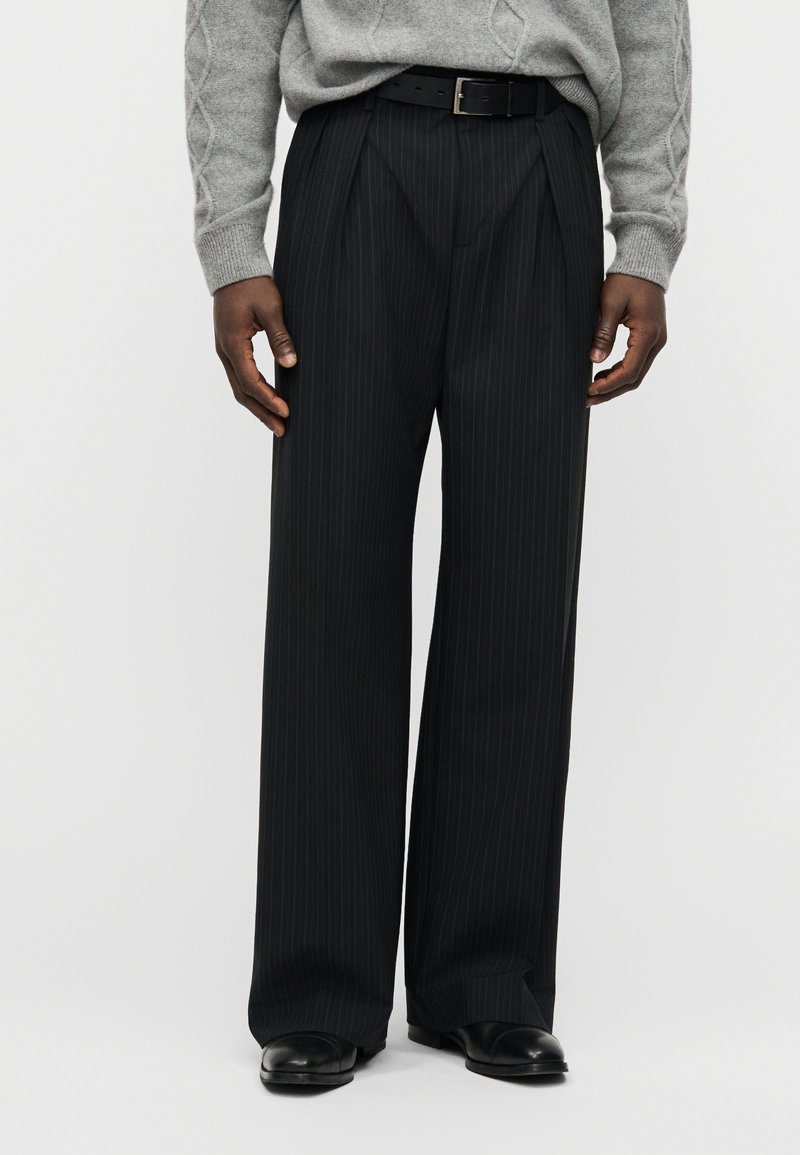 Μαύρο παντελόνι με λεπτές ρίγες κα ι άνετη εφαρμογή, με πιέτες μπροστά και απαλή υφή. Συνδυάζεται με γκρι πουλόβερ και μαύρη ζώνη.