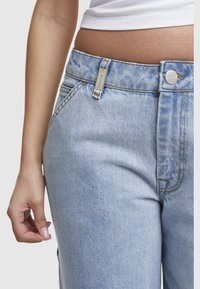 Personne portant un jean taille haute bleu clair et un crop top blanc, une main détendue sur le côté, montrant du vernis à ongles.