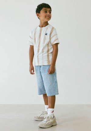 Chlapec stojí a dívá se stranou, má na sobě béžové pruhované tričko, světle modré texturované šortky, bílé ponožky a béžové tenisky.