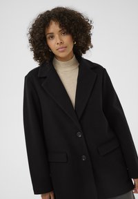 Čierny kabát z vlnenej zmesi s klopami, dvoma prednými vreckami a uzáverom na dva gombíky. Nosí sa cez smotanový rolák.