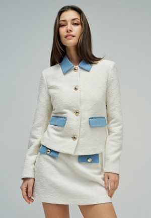 Donna con completo giacca e gonna bianco strutturato con colletto e pattine delle tasche azzurro chiaro, in piedi su sfondo grigio uniforme.