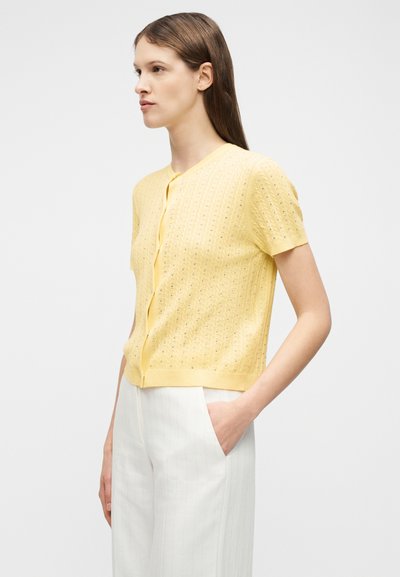 Γυναίκα που φοράει ένα κίτρινο ζακέτα με κουμπιά και κοντά μανίκια, με διακριτικό σχέδιο, και άσπρο παντελόνι ψηλόμεσο, στέκεται με το χέρι στην τσέπη.