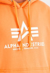 Sudadera naranja con logo blanco y el texto "ALPHA INDUSTRIES" en el pecho. Presenta cordones, bolsillo frontal y una textura de tela suave.
