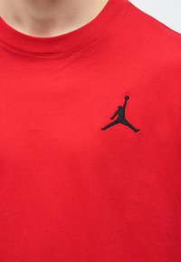Punainen puuvillainen T-paita, jossa on pyöreä kaulus ja lyhyet hihat. Vasemmassa rinnassa musta kirjailtu Jumpman-logo.