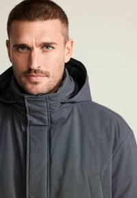 Mann mit kurzen Haaren und Bart, der eine dunkelgraue Kapuzenjacke mit Reißverschluss trägt und direkt in die Kamera schaut, vor einem schlichten Hintergrund.