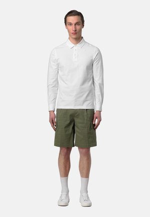 Giovane uomo in piedi immobile che indossa una polo bianca a maniche lunghe, pantaloncini cargo verde oliva, calzini bianchi e scarpe da ginnastica bianche su uno sfondo semplice.