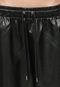 Prim-plan al pantalonilor scurți negri din piele ecologică perforată, cu talie elastică și șireturi negre cu capete metalice.