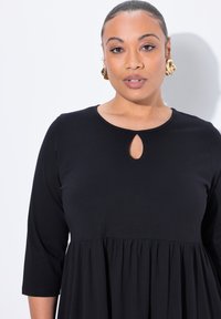 Musta mekko, jossa on kolme neljännesosaa hihat, pyöreä pääntie ja avainreikäyksityiskohta. Kangas on sileä ja siinä on kootut vyötärölinjat.