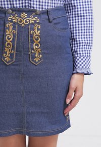 Gonna di denim con ricami dorati sulla parte anteriore, caratterizzata da vita alta, taglio dritto e dettagli di cucitura gialla. Camicia a quadri blu in vista.