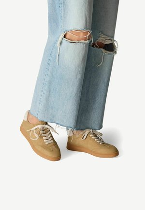 Sneaker beige in materiali scamosciati e in pelle, con punte rotonde, lacci bianchi e suola in gomma color caramello; abbinati a jeans azzurri chiari strappati.