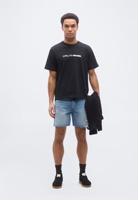 Mand iført sort Calvin Klein t-shirt, denimshorts og sorte sneakers, der holder en sort sweater mod en ensartet hvid baggrund.