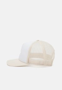 Krém és fehér trucker sapka szilárd elülső résszel, hálós hátsó résszel és ívelt karimával; tetején gomb található, és állítható snapback záródású.