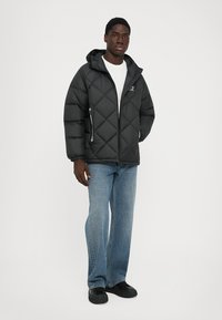 Jachetă puffer neagră cu glugă, model cu carouri în formă de diamant, cu buzunare cu fermoar în față, purtată peste un tricou alb și blugi bleu deschis. Pantofi negri.
