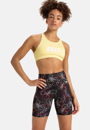 Gele sportbeha met "ROXY" logo; zwarte shorts met een kleurrijk bloemenpatroon; rekbare stof; strakke pasvorm.