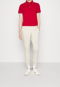 Personne portant un polo rouge à manches courtes, un pantalon beige retroussé, des chaussettes blanches et des baskets blanches avec des accents rouges, se tenant contre un fond uni.