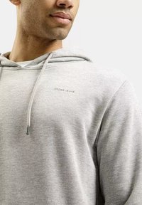 Mężczyzna w jasnoszarej bluzie z kapturem z regulowanymi sznureczkami i małym logo "CROSS JEANS" na piersi, na jednolitym jasnym tle.