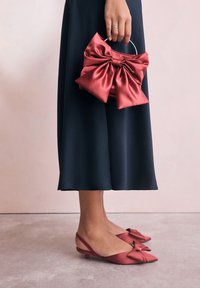 Rdeče slingback čevlje iz satena z detajlom pentlje, kombinirane z usklajeno rdečo satinasto torbico z mašno in srebrno ročico, postavljene ob temno krilo.