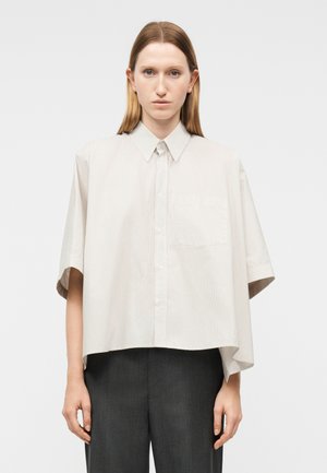 Ženska, oblečena v ohlapno, svetlo bež srajco s kratkimi rokavi, gumbi spredaj in žepom na prsnem delu ter temno sive hlače, stoji pred enostavnim ozadjem.