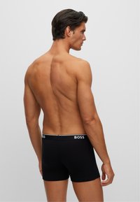 Schwarze Boxershorts mit einem Logoschlüpfer, aus einem glatten, dehnbaren Material gefertigt. Die Rückansicht zeigt ein eng anliegendes Design und einen leichten Glanz.