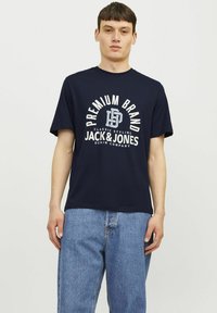 Tamno plava pamučna majica s bijelim grafičkim otiskom koji prikazuje tekst "PREMIUM BRAND" i "JACK & JONES" na prednjoj strani. Kratki rukavi.