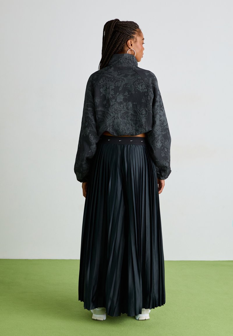 Chaqueta bomber negra corta con un patrón floral texturizado, combinada con una falda maxi negra plisada y zapatillas blancas, vista desde atrás.