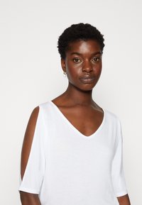 Witte T-shirt met korte mouwen, een V-hals en uitsnijdingen op de schouders, gemaakt van lichte stof, met een gladde textuur en een ontspannen pasvorm.