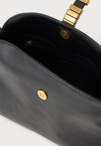 Crna kožna torbica s zlatnim magnetskim zatvaračem i lančićem, djelomično otvorena, prikazuje crni unutrašnjost s džepom na zip.