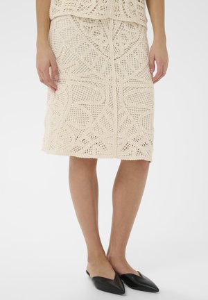 Gebreide kokerrok in crème met ingewikkelde openwerkontwerpen en geometrische patronen, gecombineerd met zwarte puntige instapschoenen.