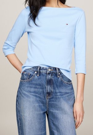 Frau trägt ein hellblaues Dreiviertelarm-Top, das in eine hoch taillierte, verwaschene blaue Jeans gesteckt ist, eine Hand in der Tasche, vor einfarbigem Hintergrund.