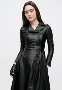 Μαύρο δερμάτινο φόρεμα με εφαρμοστό μπούστο και φαρδιά φούστα, με γιακά, κλείσιμο με φερμουάρ και πλαϊνές τσέπες. Γάντι στο δεξί χέρι.