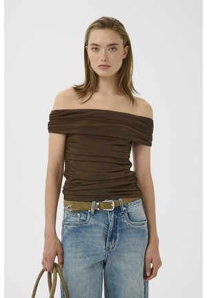 Junge Frau trägt ein braunes schulterfreies, gerafftes Oberteil, hellblaue Jeans mit einem braunen Gürtel und hält eine passende braune Handtasche vor weißem Hintergrund.