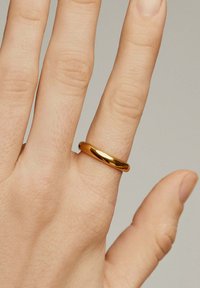 Anillo de metal color dorado con una banda lisa y redondeada, llevado en una mano que muestra los dedos. La superficie tiene un acabado reflectante.