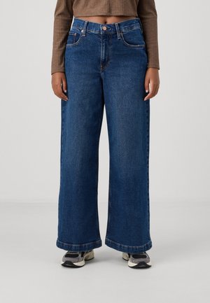 Person iført blå jeans med vide ben, brun kort sweater og grå sneakers, stående mod en ensfarvet lysegrå baggrund.