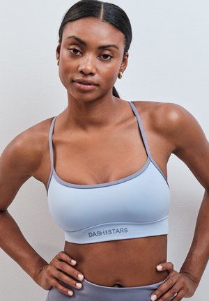 Femme aux cheveux foncés portant un brassière de sport bleu clair de la marque "Dash and Stars", debout les mains sur les hanches contre un mur gris.