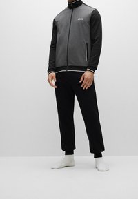 Giacca grigia con zip, maniche nere, dettagli bianchi e un piccolo logo. Abbinata a pantaloni neri e calze bianche. Vestibilità slim, tessuto morbido.