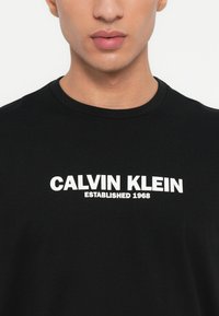Melnas kokvilnas T-krekls ar apaļu kakla izgriezumu, uz krūtīm ar baltiem drukātiem vārdiem "CALVIN KLEIN ESTABLISHED 1968". Gluda tekstūra.