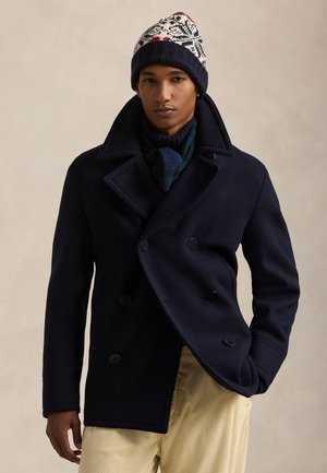 Homme portant un caban bleu marine, un pantalon en velours côtelé crème, un bonnet tricoté à motifs et une écharpe à carreaux foncée sur un fond beige uni.