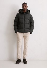 Ciemnozielona pikowana kurtka z kapturem, zamkiem błyskawicznym i puchatym fasonem, zestawiona z jasnobeżowymi spodniami i ciemnymi butami. Gładka tekstura.